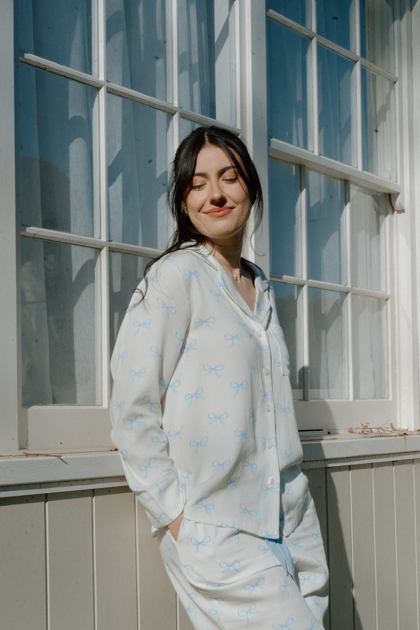 Young woman leaning against a window outside, wearing a white long sleeve pyjama set with a blue bow print